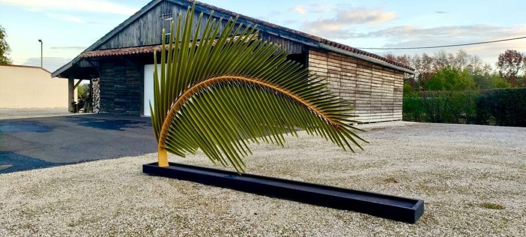 Sculpture en métal en forme de feuille de palmier arquée, posée sur un long socle noir devant un bâtiment en bois.