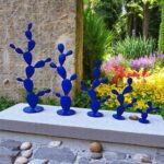 Sculptures bleues en métal en forme de cactus exposées dans un jardin coloré avec fleurs et verdure