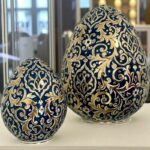 Deux œufs décoratifs bleus et dorés avec motifs floraux exposés sur une surface blanche.