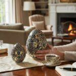 Deux œufs décoratifs bleus et dorés sur une table, avec une tasse de café et un feu de cheminée en arrière-plan.