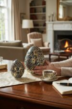 Deux œufs décoratifs bleus et dorés sur une table, avec une tasse de café et un feu de cheminée en arrière-plan.